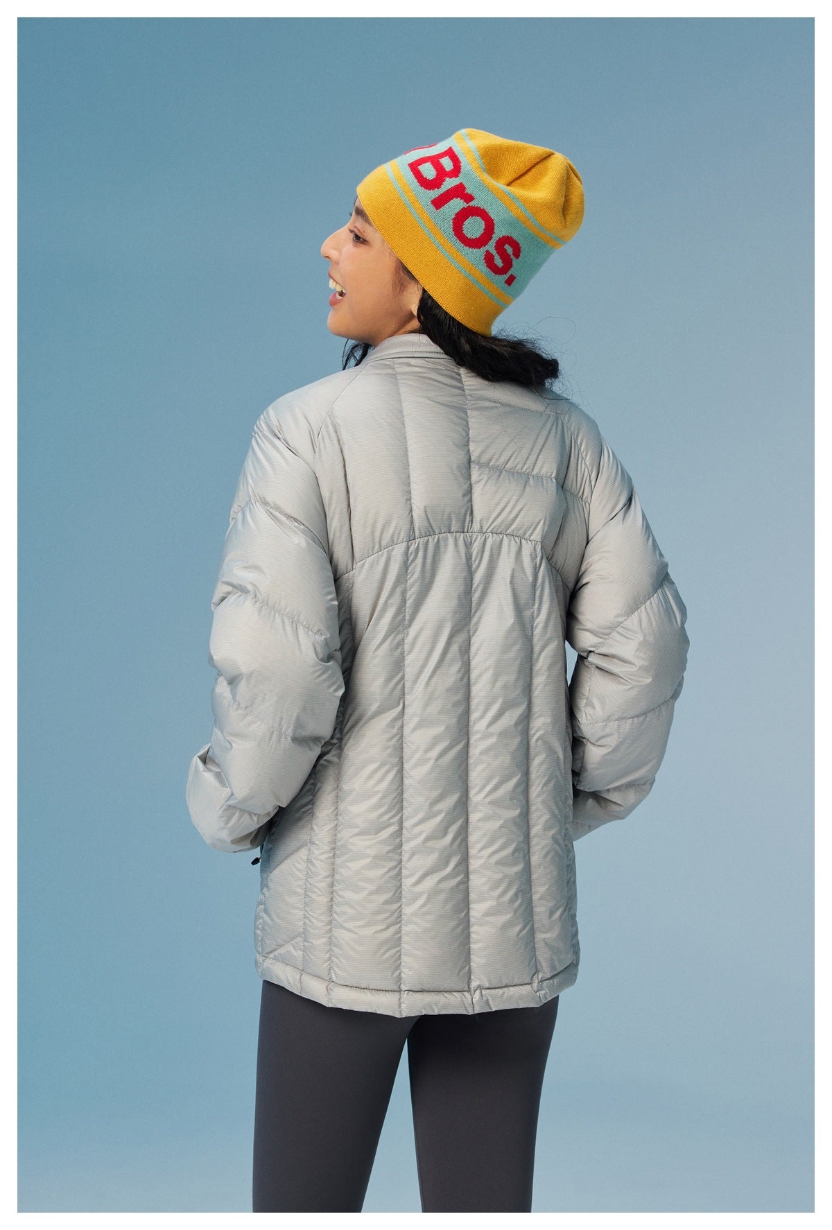 Back view of a model wearing a light gray quilted jacket and a yellow knit beanie.