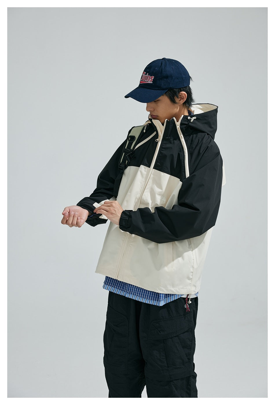 Model adjusts the cuff of a cream-and-black hooded jacket with a front zipper and contrast panels.