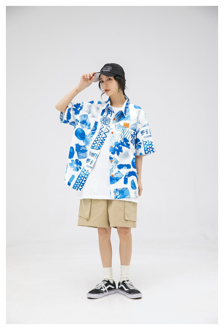 Model in blue abstract print short-sleeve shirt worn open over a white tee, adjusting a black cap