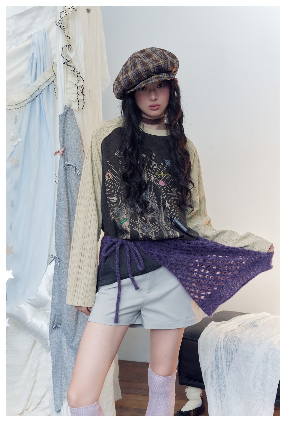 Model wearing a charcoal castle-print raglan top with cream striped sleeves, pale shorts, and a purple knit wrap.