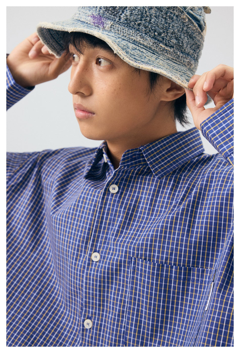 Close-up of the blue checked shirt's collar, white buttons, and chest pocket as the model adjusts a bucket hat.