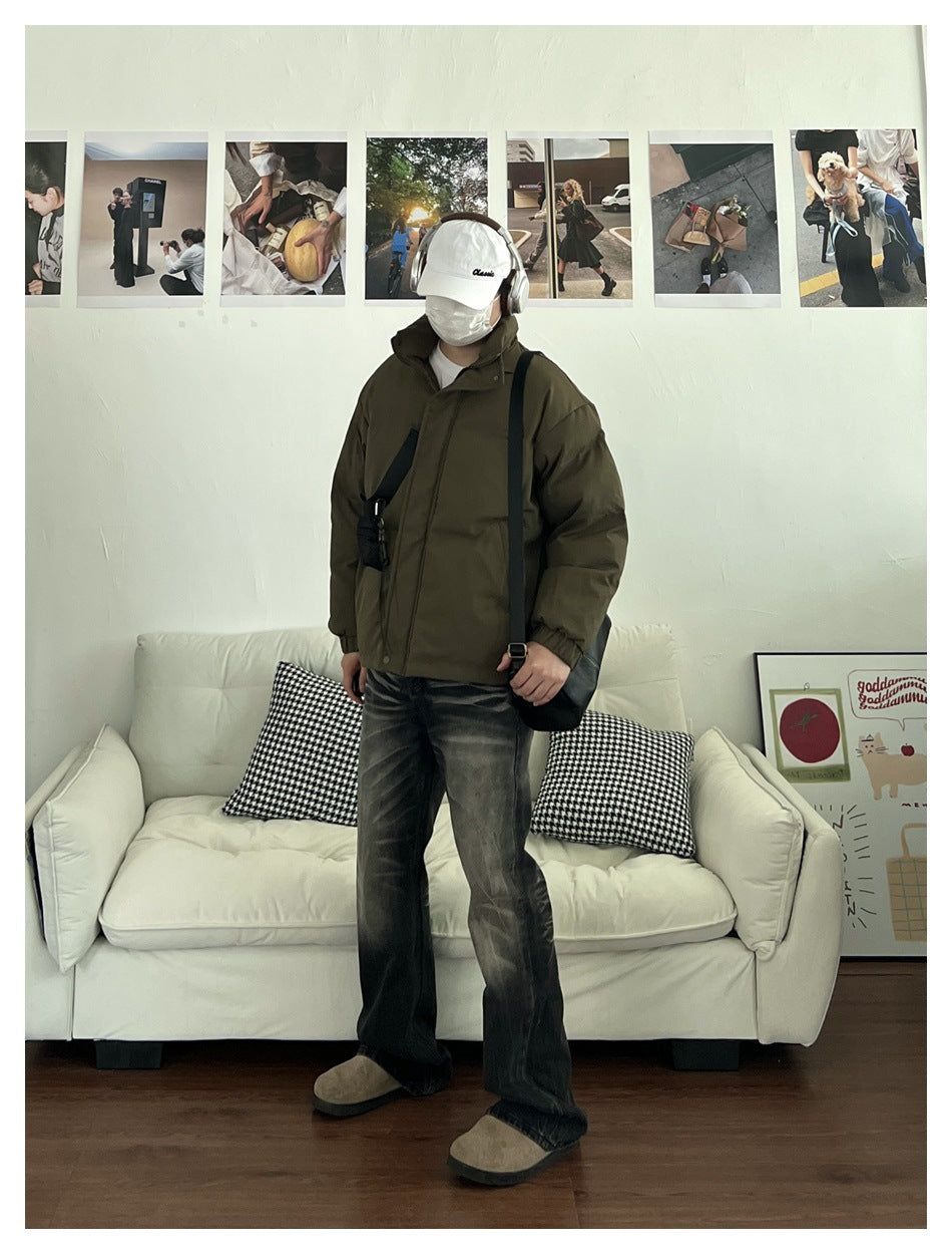 Person wearing an olive padded jacket with a high collar, dark jeans and a white cap indoors.