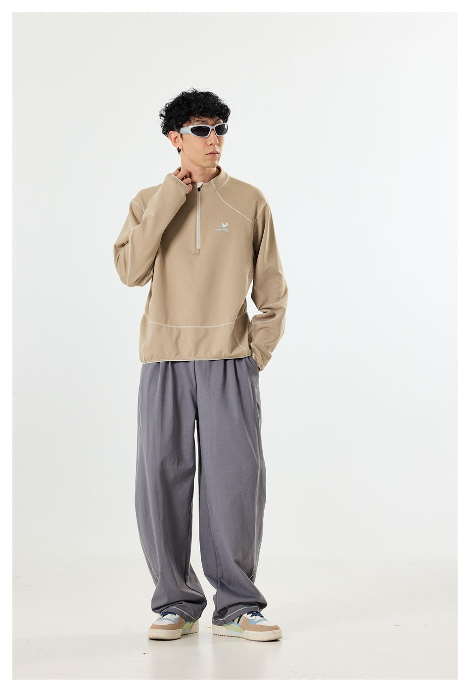A man models a beige quarter-zip pullover with pale contrast seams and loose gray pants, facing slightly to the side.