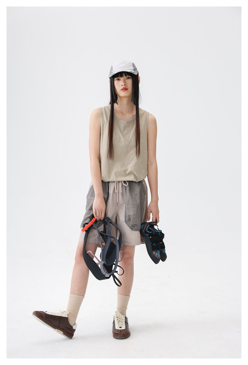 Model wearing a sand beige sleeveless tank top with shorts and a cap, holding a harness and climbing shoes