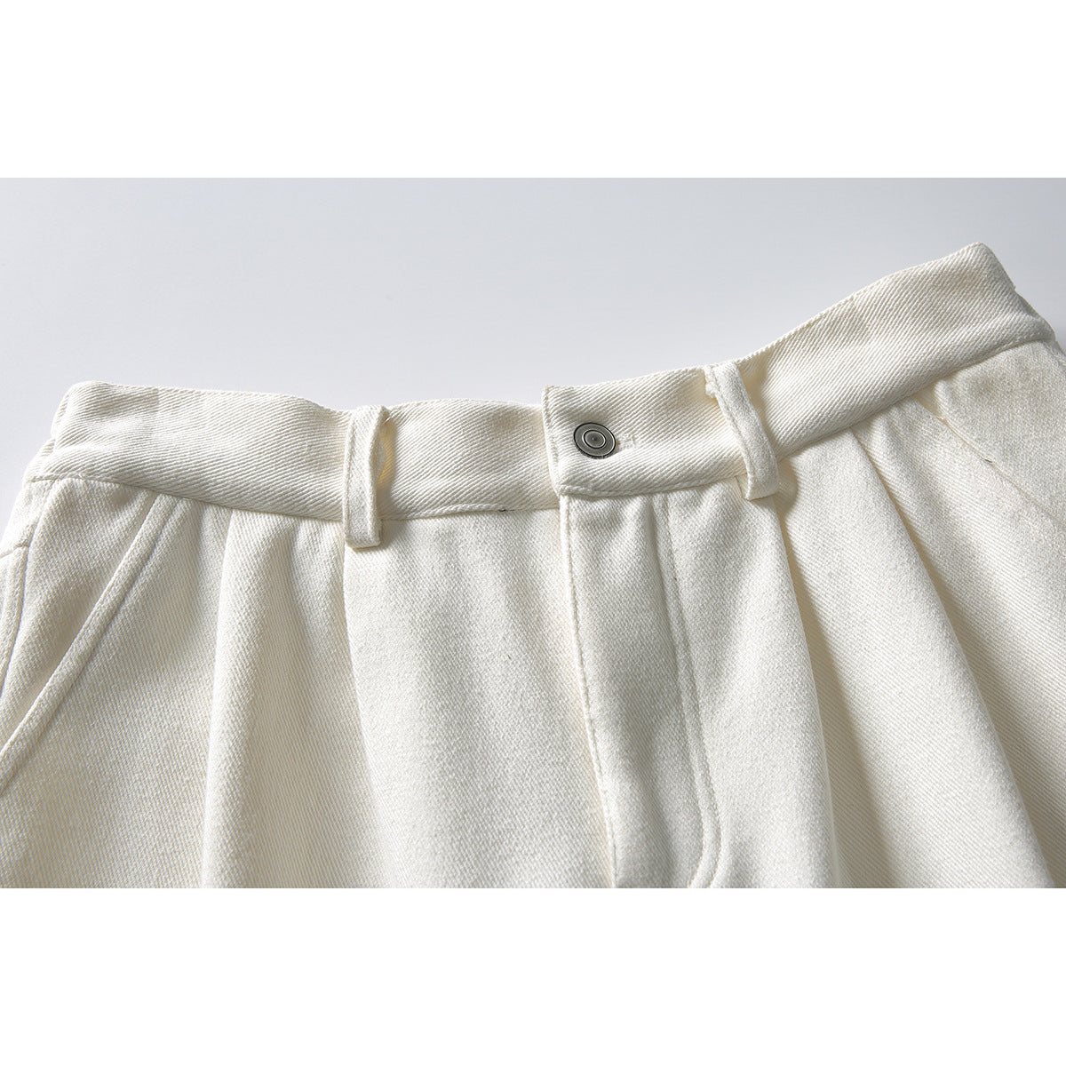 Close-up of off-white trousers showing the waistband, belt loops, metal button, pleats and pocket edge.