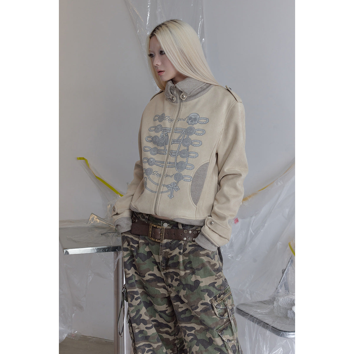 Model wearing a beige graphic zip jacket with gray ribbed trim and loose green camouflage cargo pants.