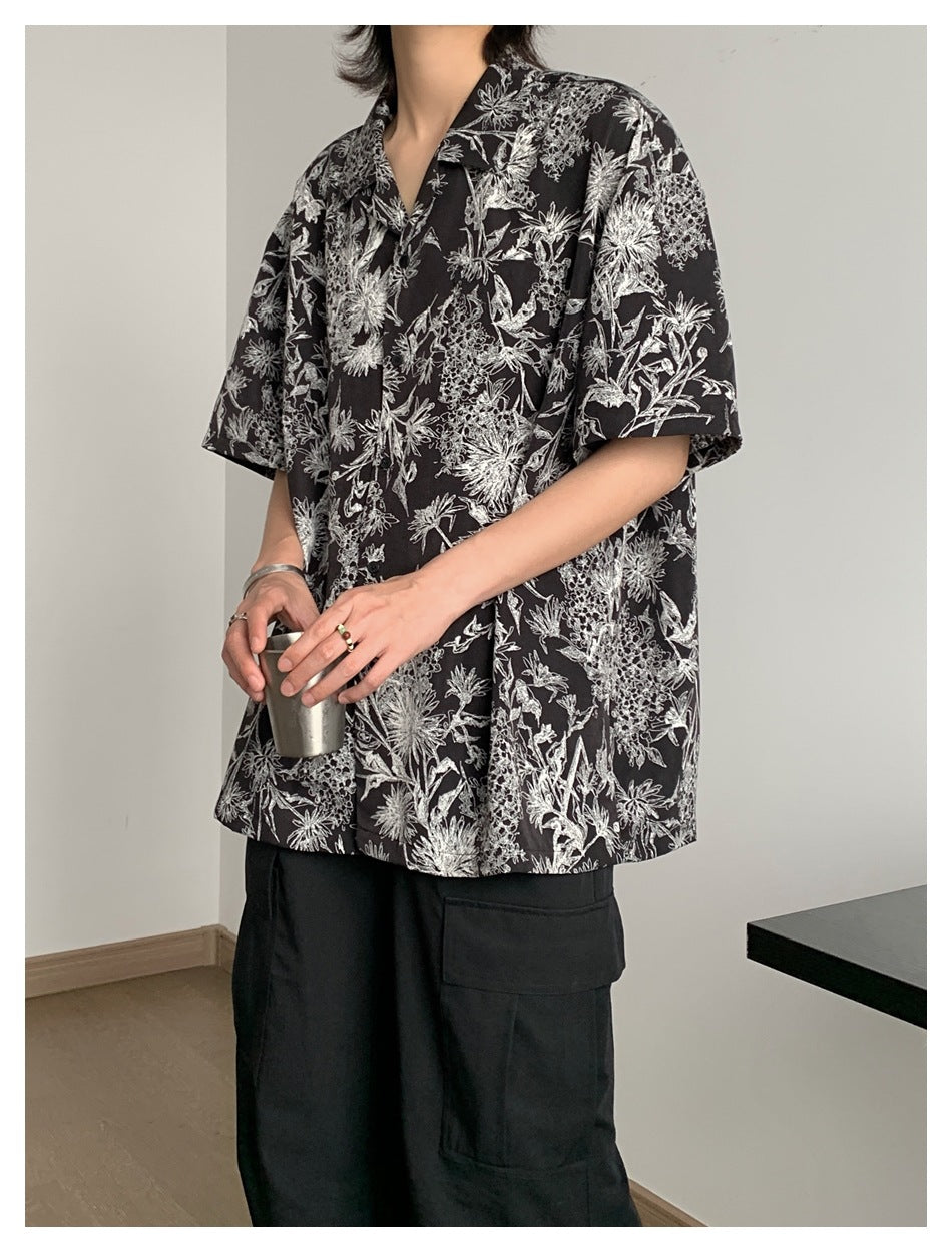 Person wearing a black short-sleeve floral-print camp-collar shirt with black cargo pants, holding a metal cup