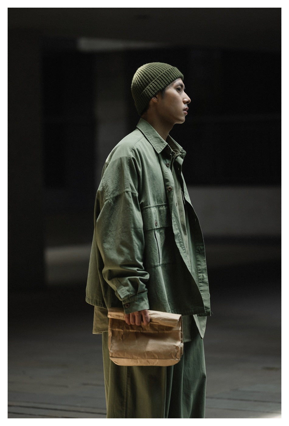 Man in profile wearing an oversized army green shirt-jacket over a green shirt, beanie and trousers, holding a paper bag