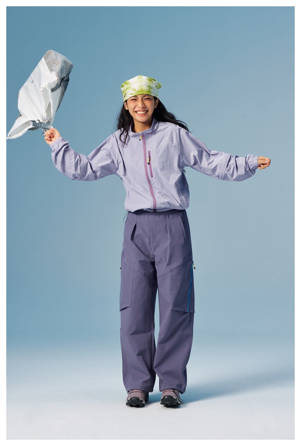 Full-length model in a pale lavender zip jacket and slate-purple cargo pants, holding a white garment against a blue
