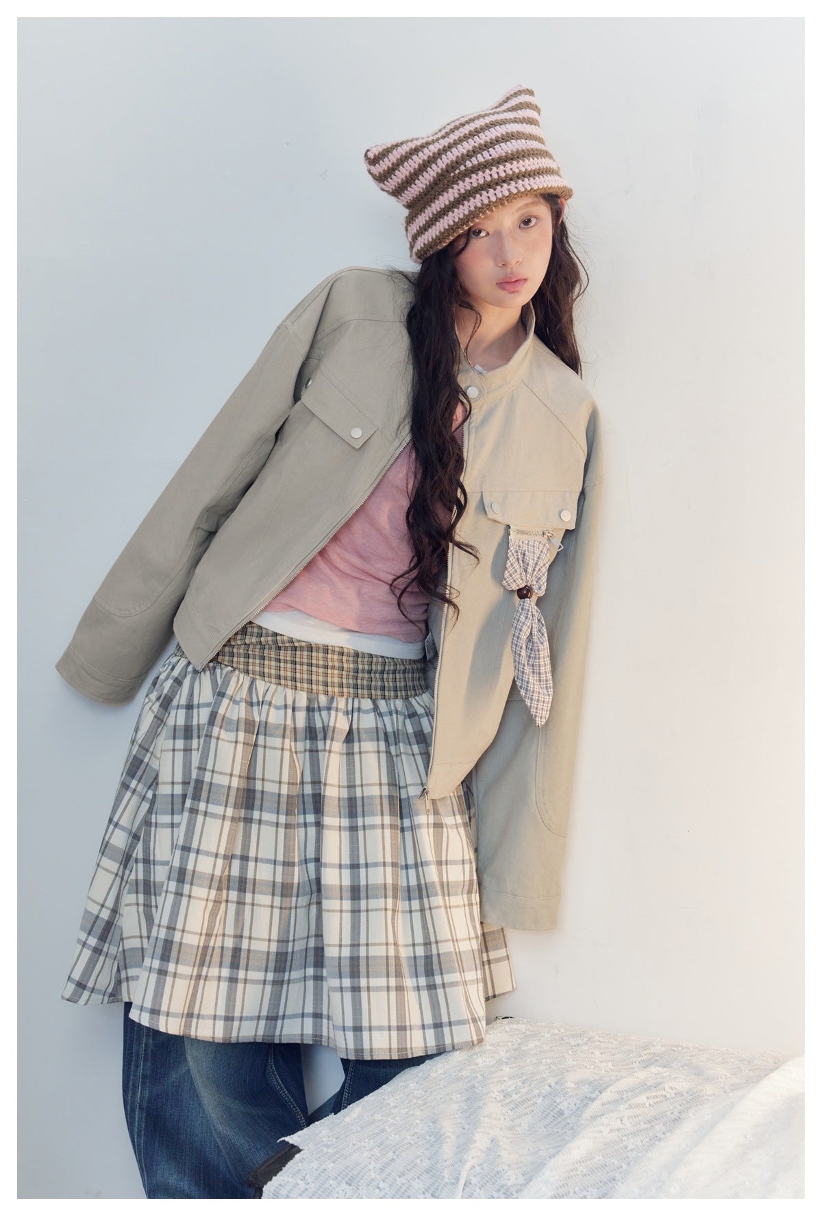 Model wearing an open light gray-beige utility jacket with snap-flap pockets and a plaid hanging detail.