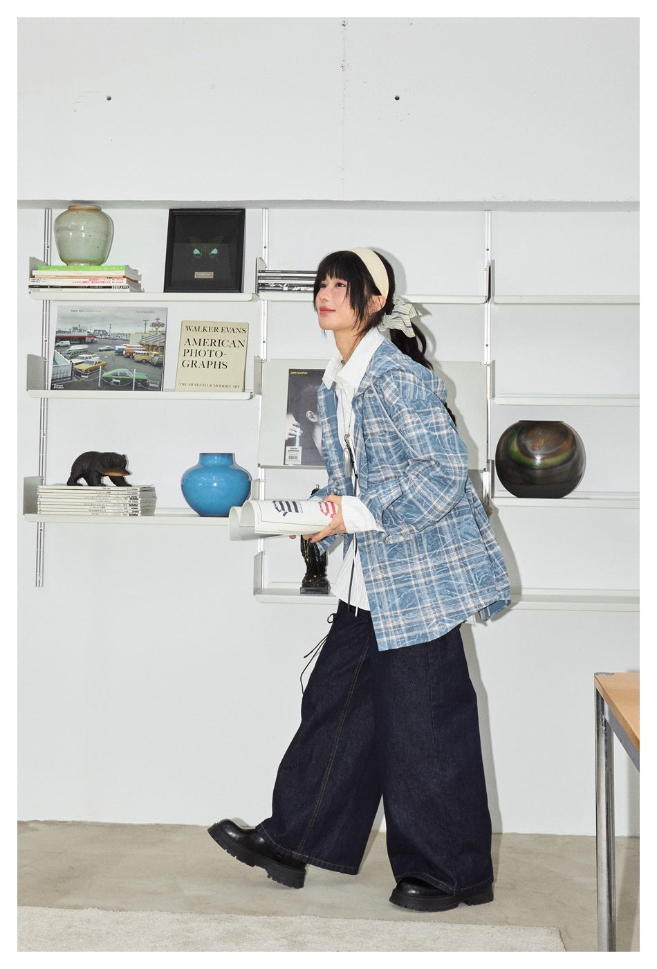 Model walking in a blue plaid hooded shirt jacket, white shirt and wide-leg dark jeans in a shelf-lined room