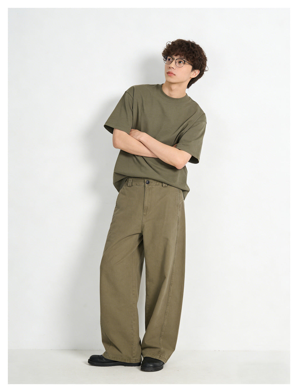 Model wearing an olive short-sleeve T-shirt and loose olive-brown trousers, standing with arms crossed.