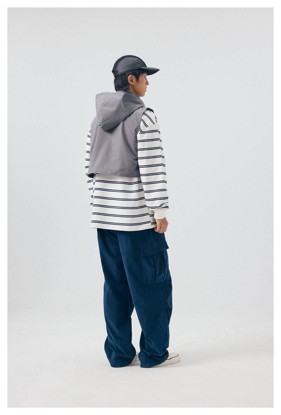 Rear view of a model wearing the gray hooded vest over a striped shirt and navy cargo pants.