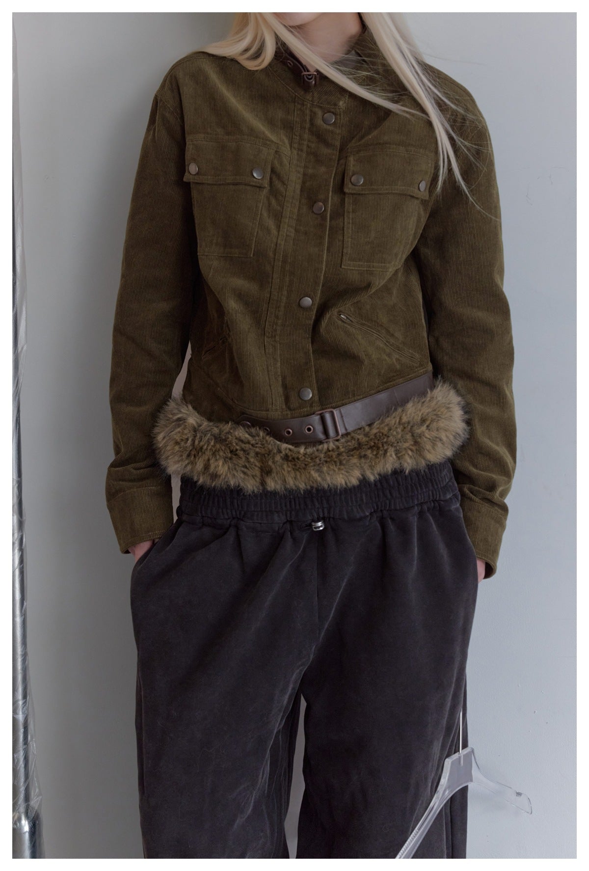 Model wearing an olive-brown corduroy jacket with snap-button chest pockets and a brown belt above a fur-trimmed waistband.