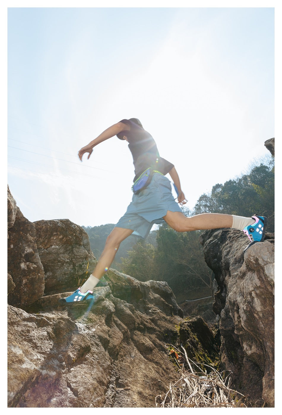 Backlit model leaping across rocks in a dark T-shirt, light blue shorts, and waist bag.