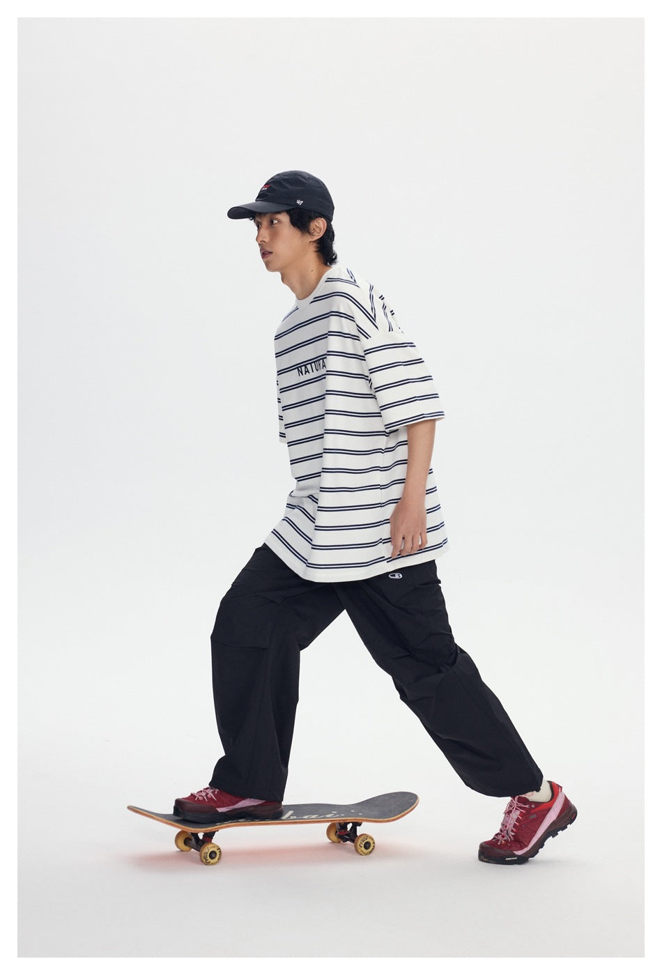 Side-view model in a white T-shirt with thin navy stripes and NATURAL text, loose black pants, and a black cap