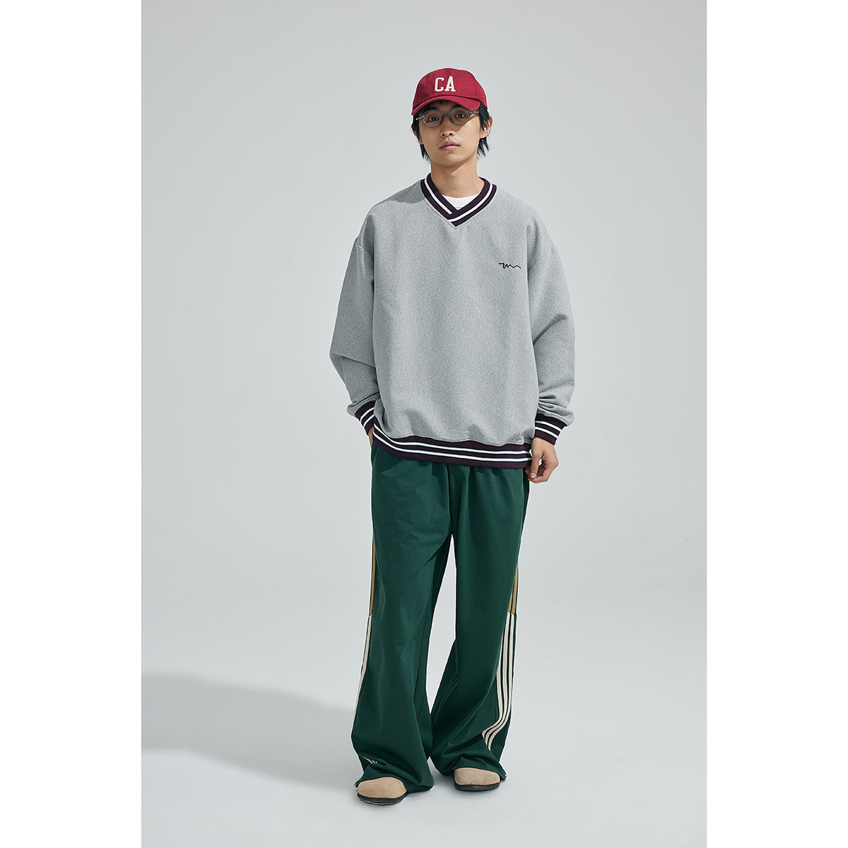 Model wearing an oversized gray V-neck sweatshirt with dark striped trim and loose green track pants.