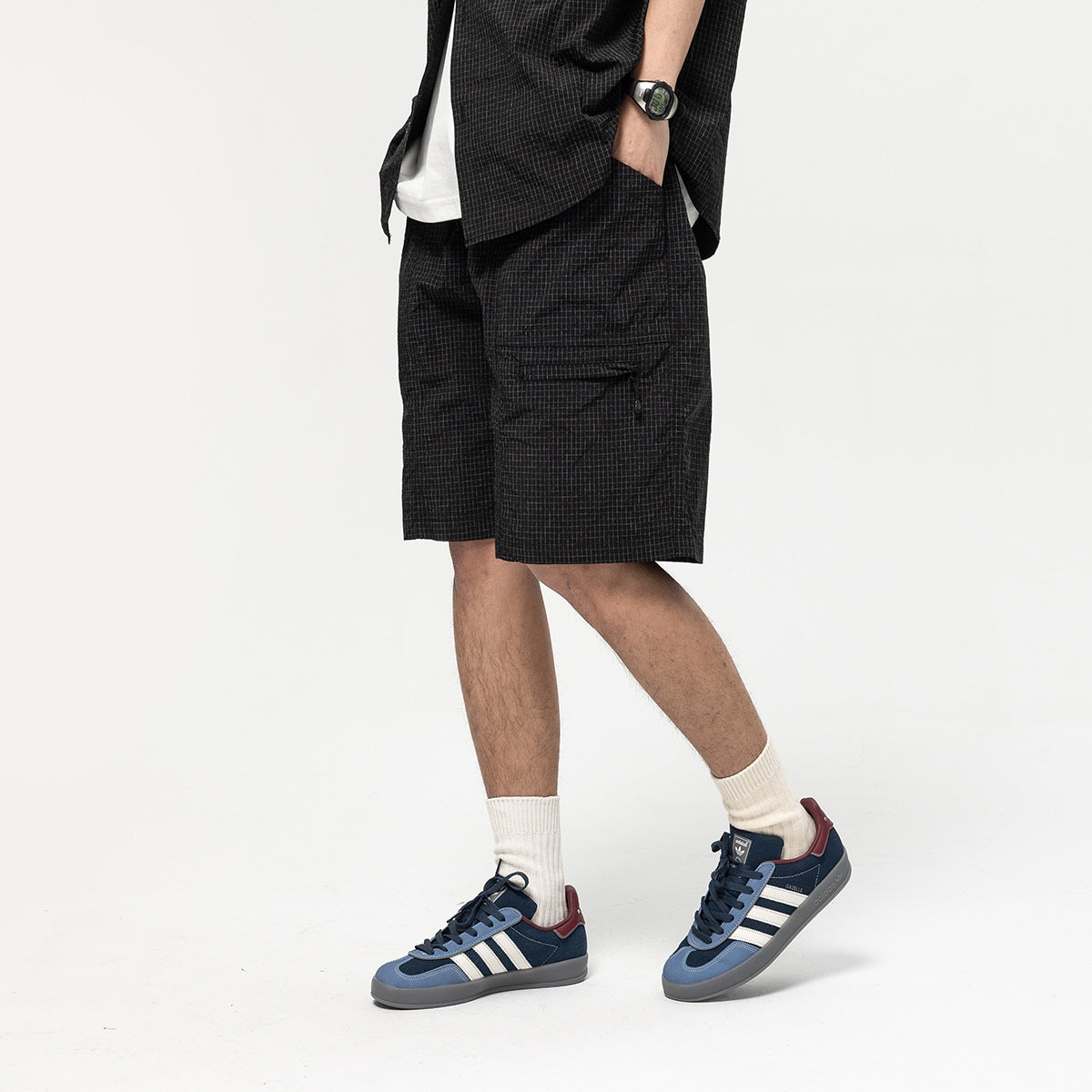Model wearing black grid-pattern knee-length shorts with a zip thigh pocket, hand in pocket, with navy sneakers