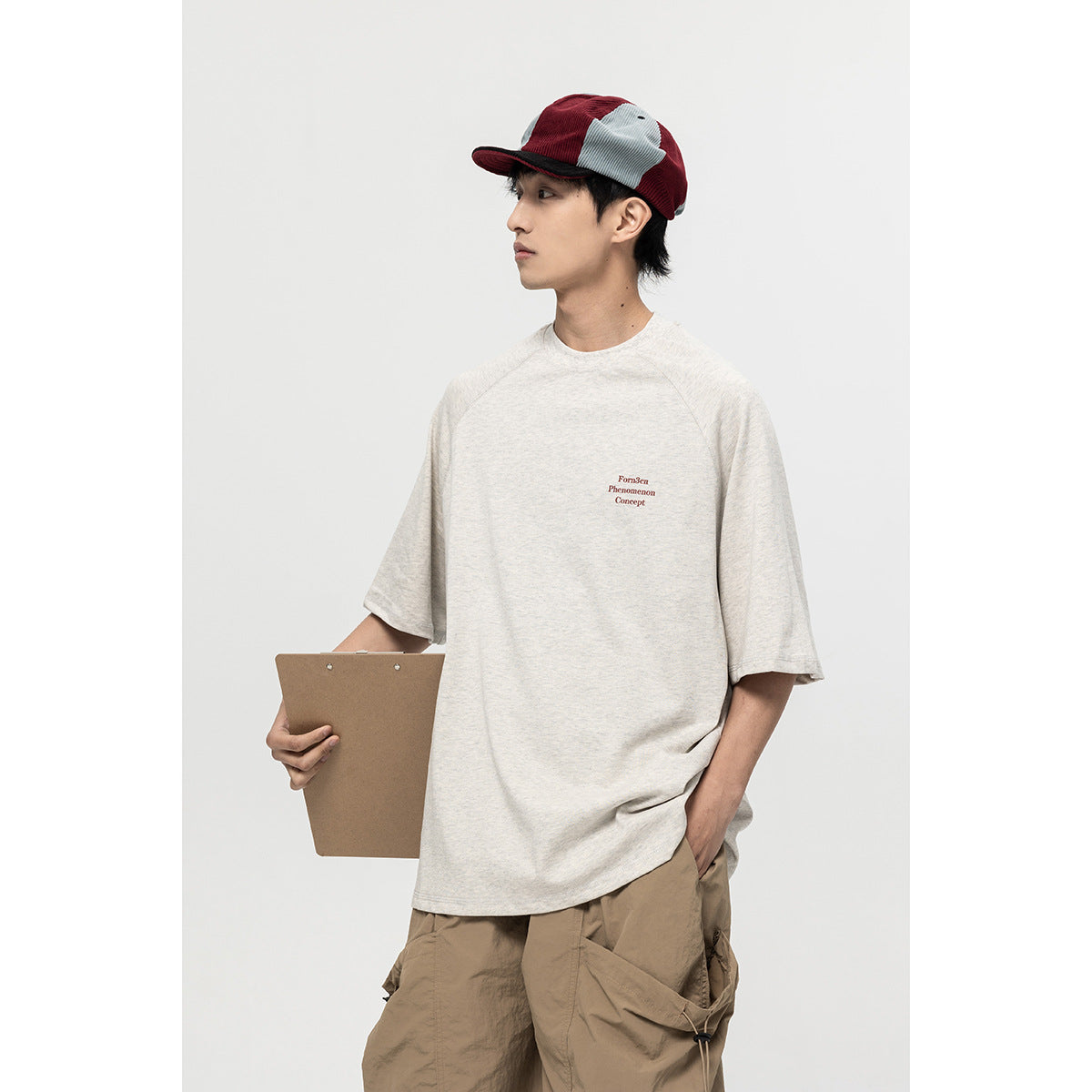 Model wearing an oversized off-white T-shirt with small burgundy chest lettering and a red-and-gray cap.