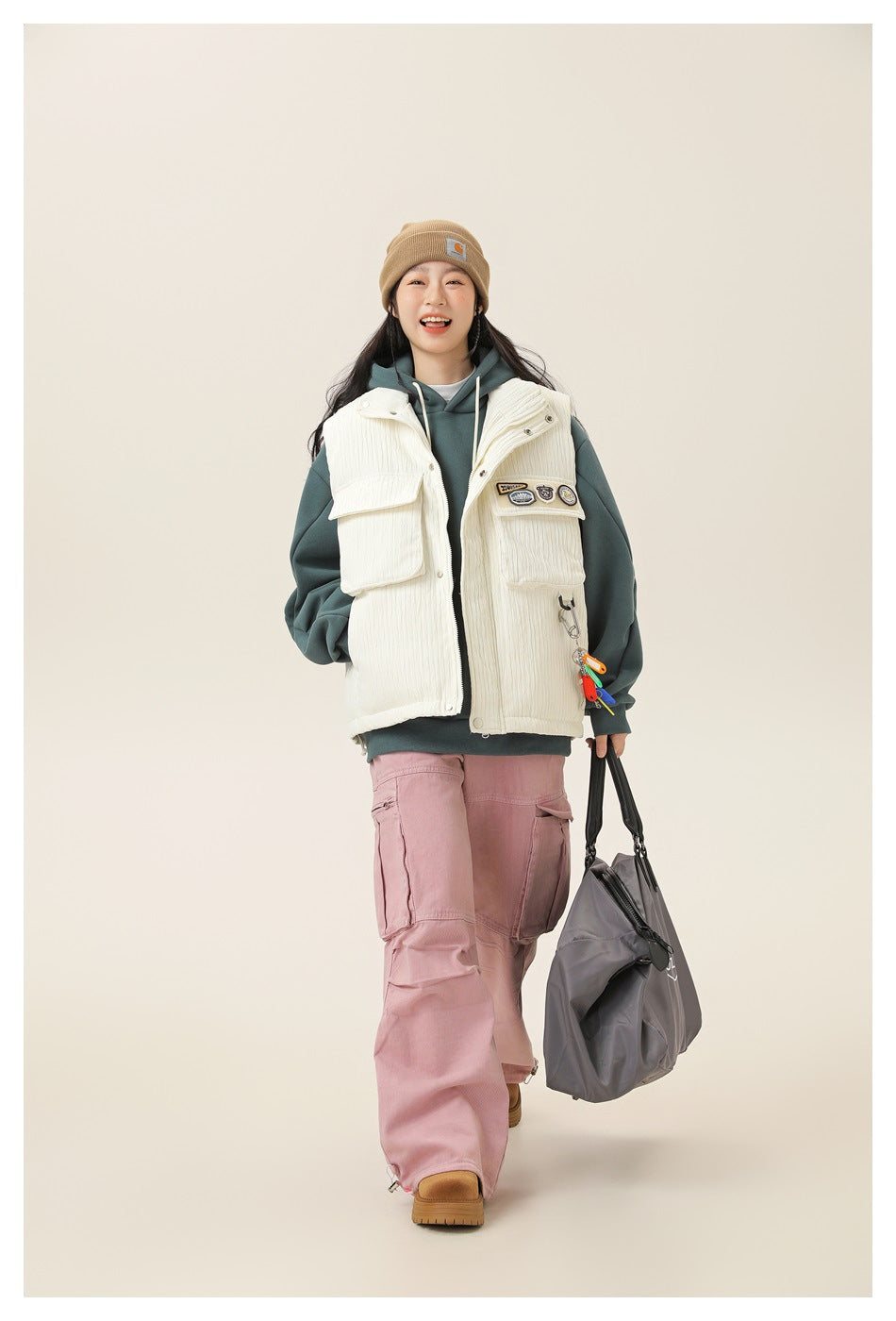 Model walks wearing an ivory utility vest over a teal hoodie and pink cargo pants, carrying a gray tote.