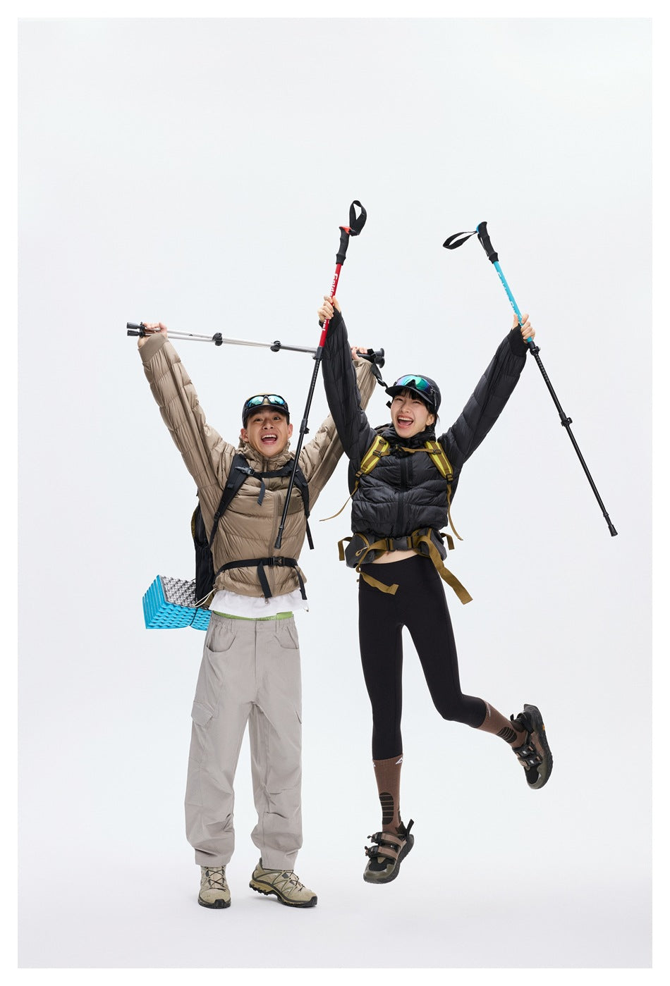 Two hikers in taupe and black hooded puffer jackets raise trekking poles against a white studio background.