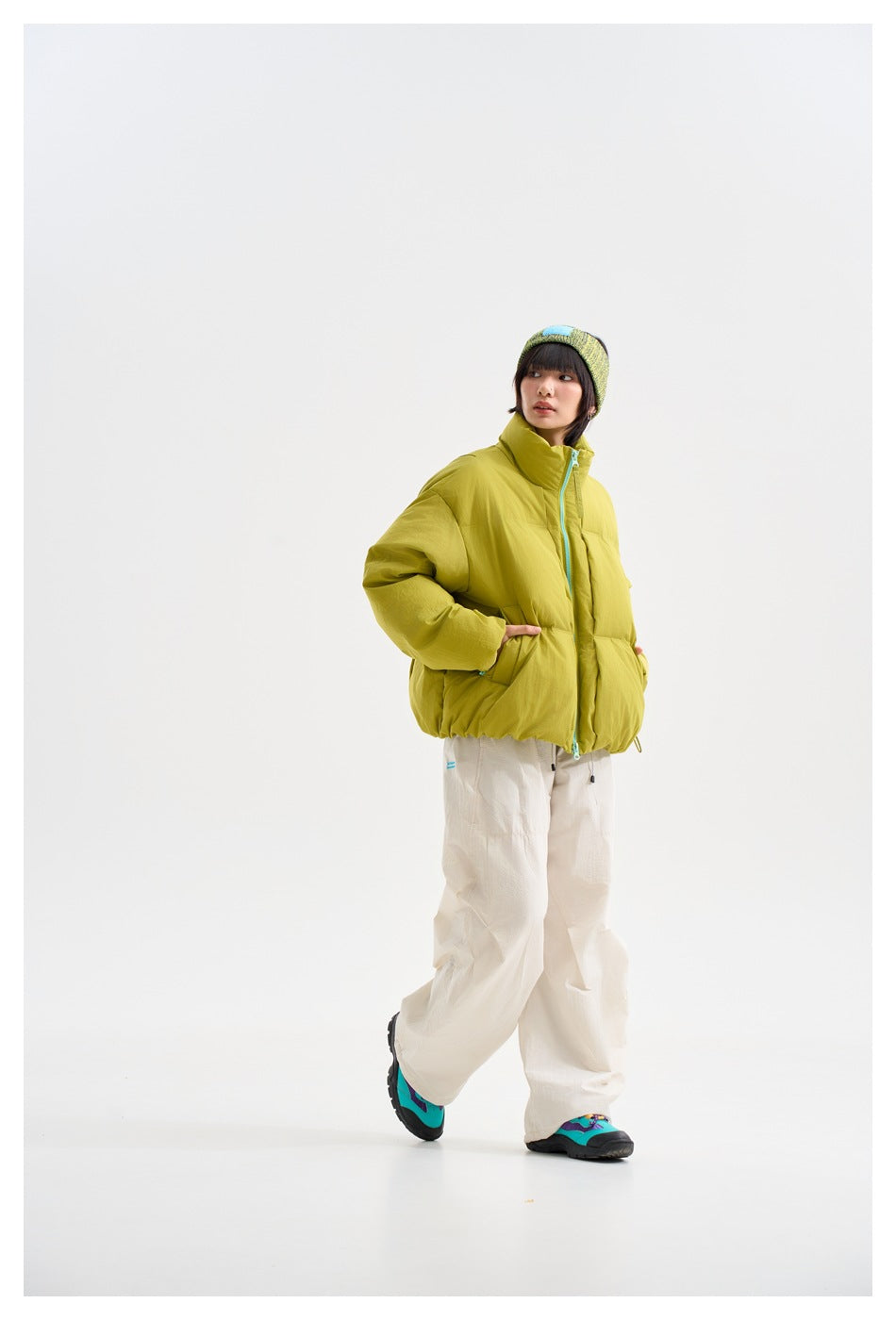 Model in green puffer jacket with hands in pockets, wearing cream pants, a knit headband and teal shoes