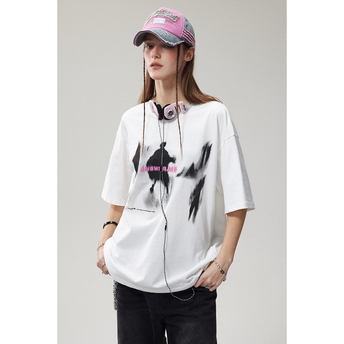 Model wearing an oversized white graphic T-shirt with black pants, a pink cap, and pink headphones.