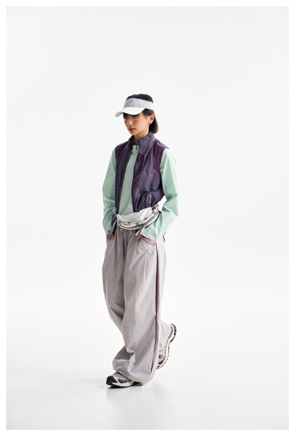 Three-quarter view of a woman in a plum utility vest, mint top, loose gray pants, white visor and waist pack.