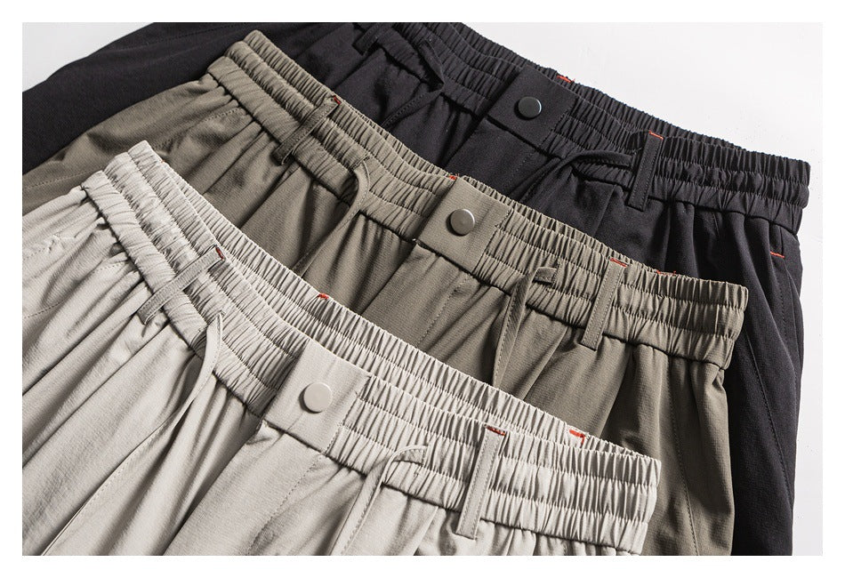 Three pairs of trousers in light gray, taupe, and black, overlapping to show elastic waistbands, buttons, and pockets.