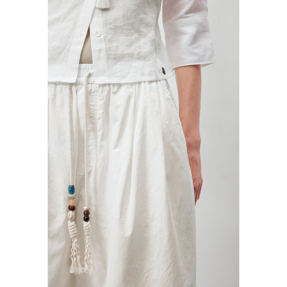 Close view of white drawstring trousers with colorful beads and tassels, worn with a white button-front top.