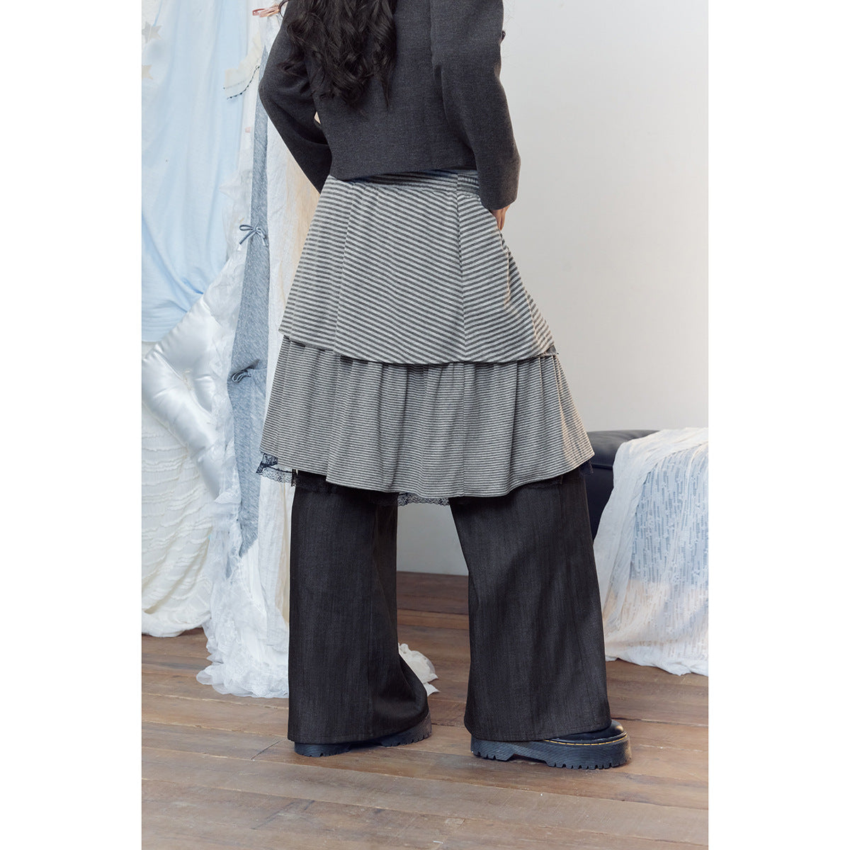 Back view of a model wearing a gray striped layered skirt over wide charcoal pants and a dark jacket.