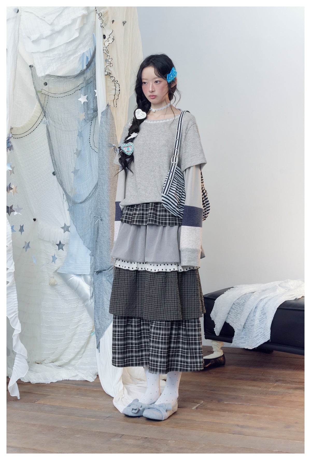 Full-length model wearing a gray layered-sleeve top and long tiered dark plaid skirt with a striped bag.