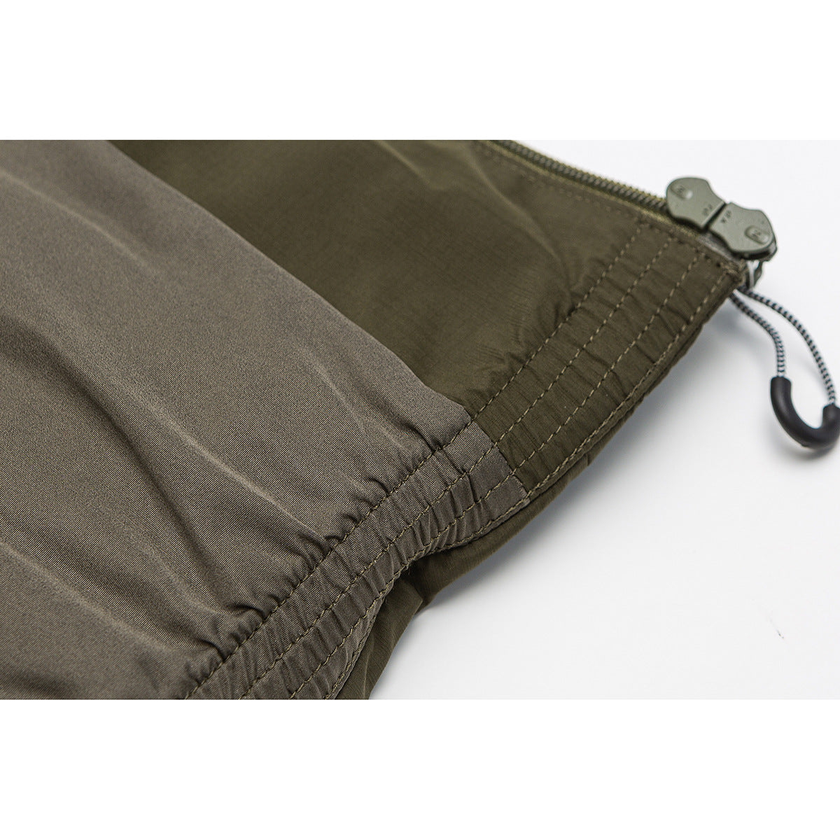 Close-up of olive garment fabric, a stitched edge and a zipper with an adjustable cord pull.