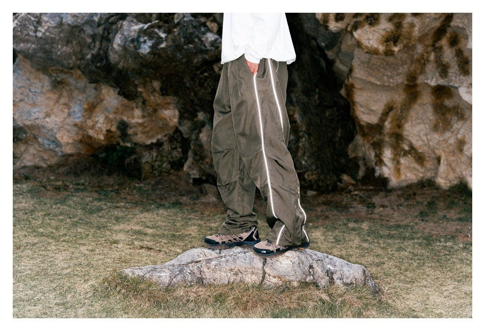 Side view of a model wearing loose taupe-gray pants with reflective piping along the outer leg.