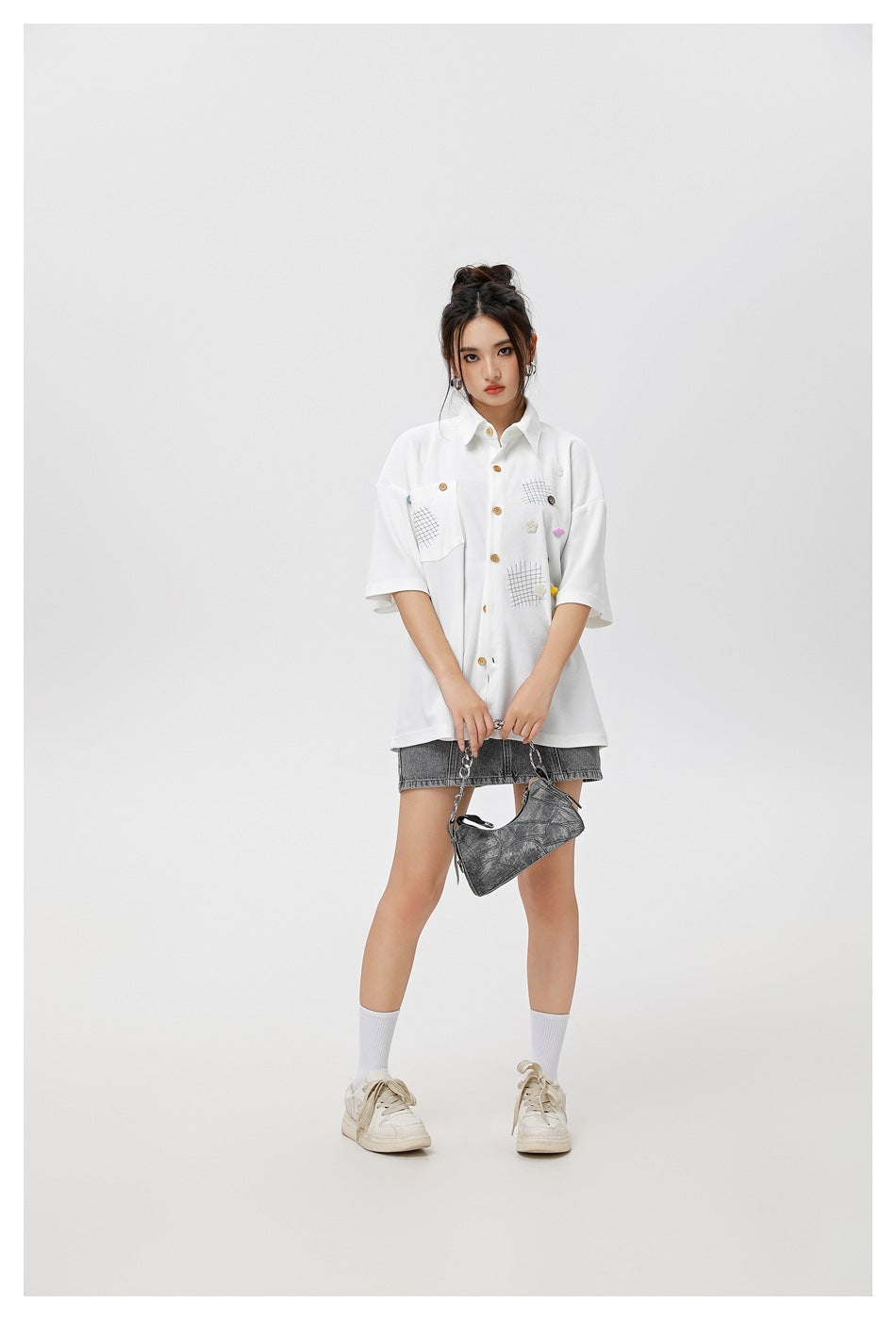 Model wearing a white oversized short-sleeve button-up shirt with a chest pocket, grid patches and colorful accents.