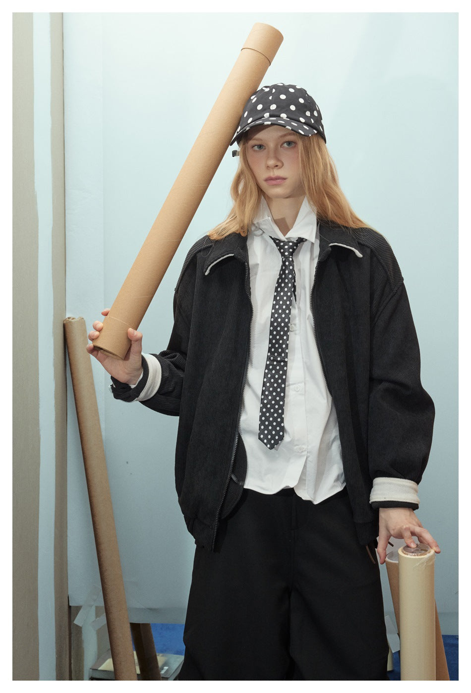 Front view of a model in an oversized black jacket with cream trim, white shirt and polka-dot cap and tie.