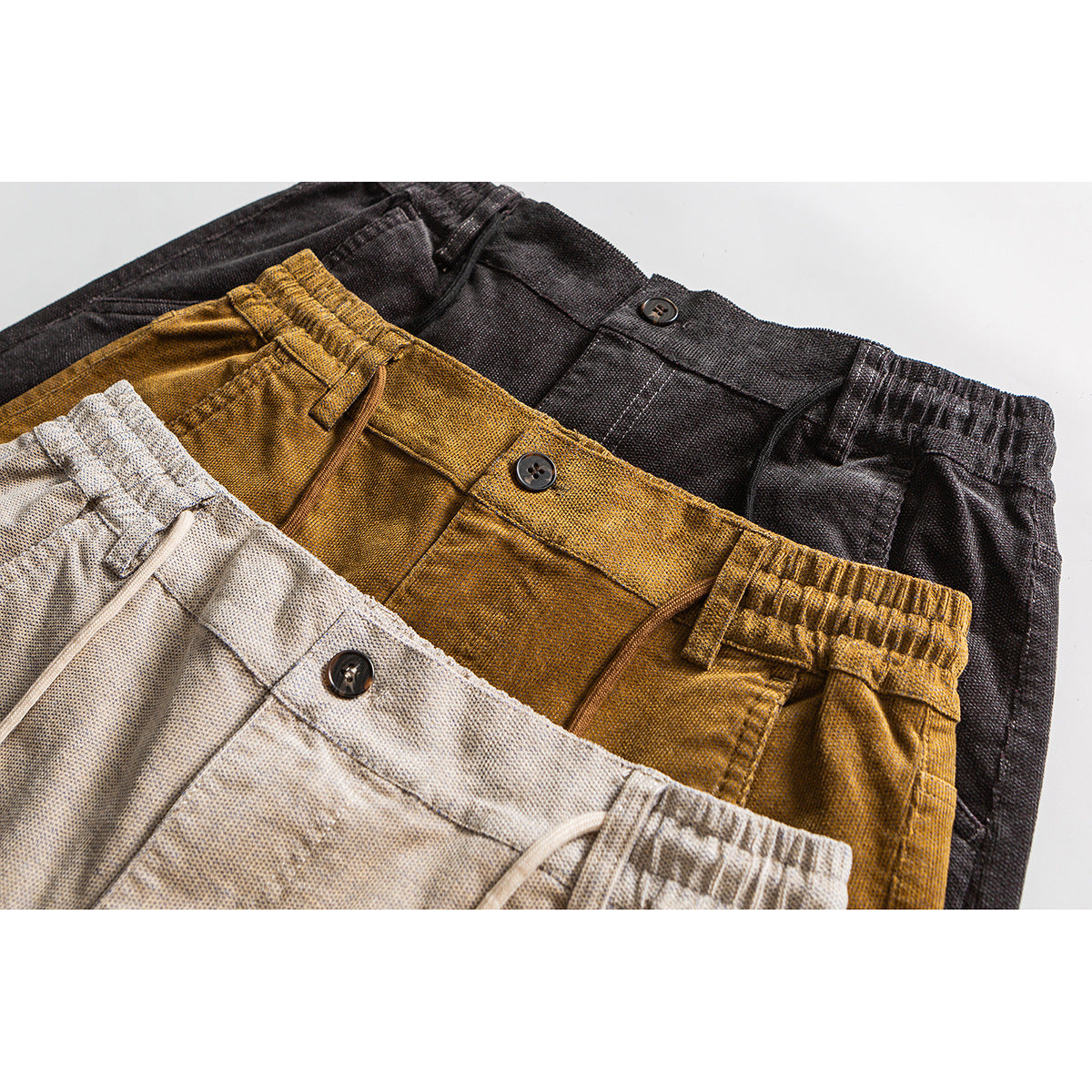 Close-up of three pairs of drawstring pants in beige, ochre, and charcoal, showing elastic waists and front pockets.