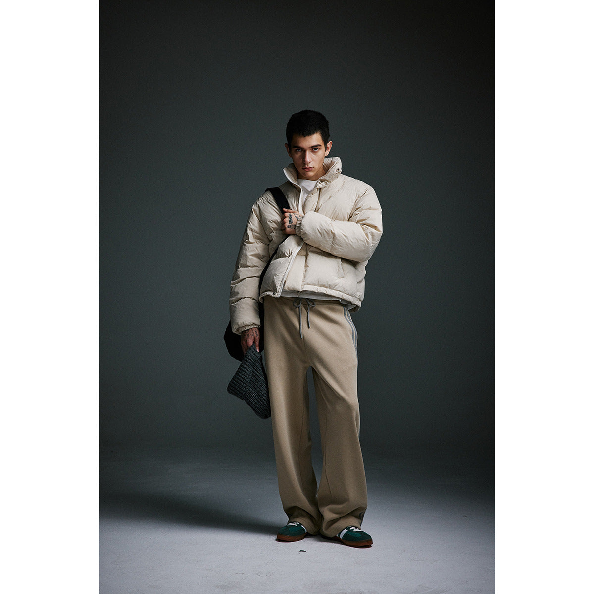 Model wearing an ivory high-collar puffer jacket and loose taupe trousers, holding a dark knitted bag.