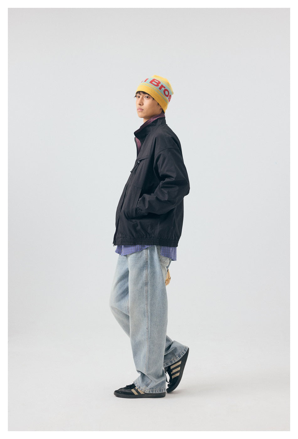 Full-length side view of a model wearing a black windbreaker, checked shirt, and loose light jeans.