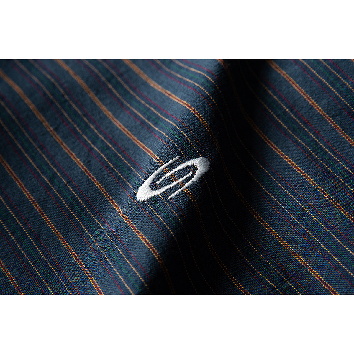 Close-up of a small white embroidered oval emblem on dark blue fabric with multicolored pinstripes.