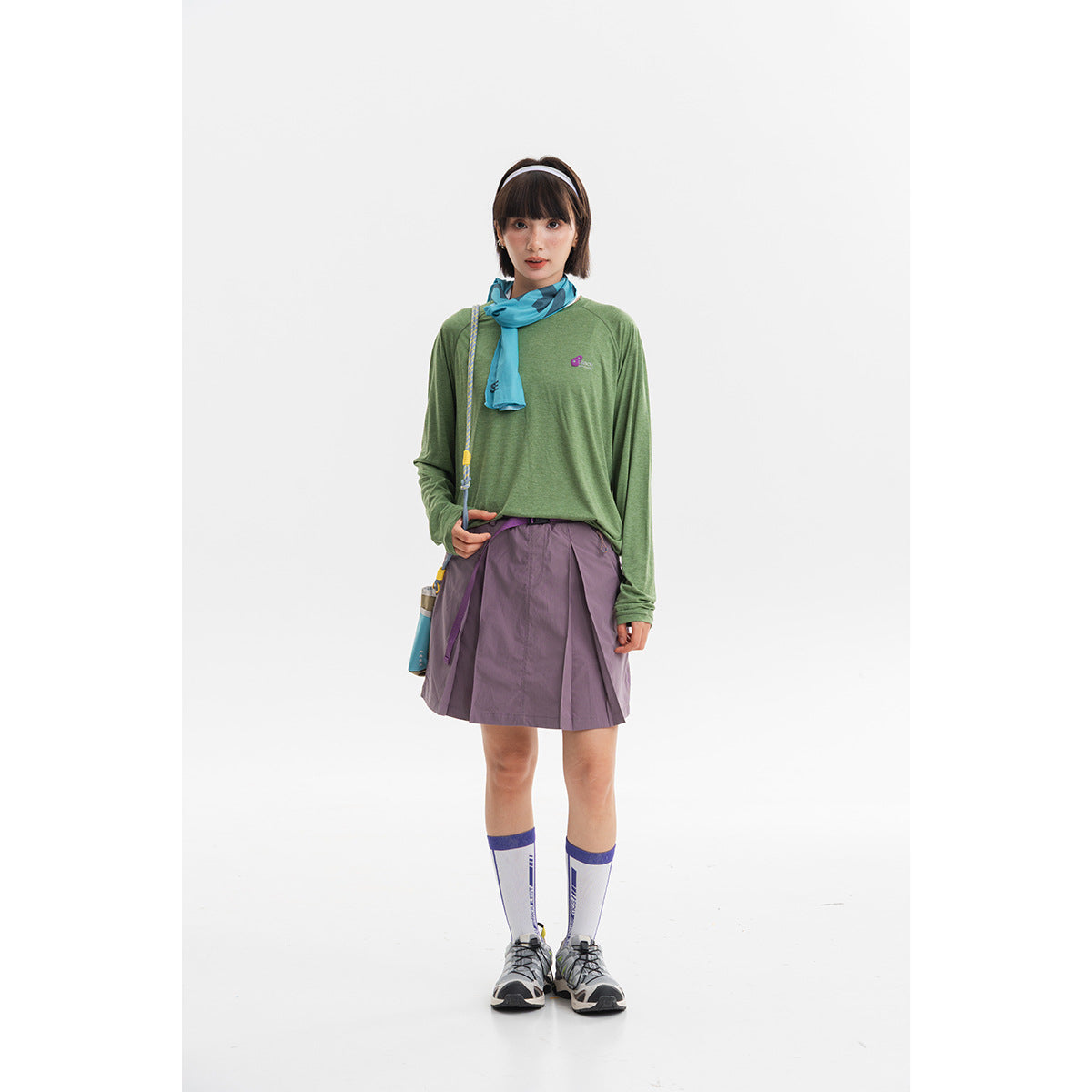 Model wearing a green long-sleeve top, turquoise scarf, and purple pleated skirt.