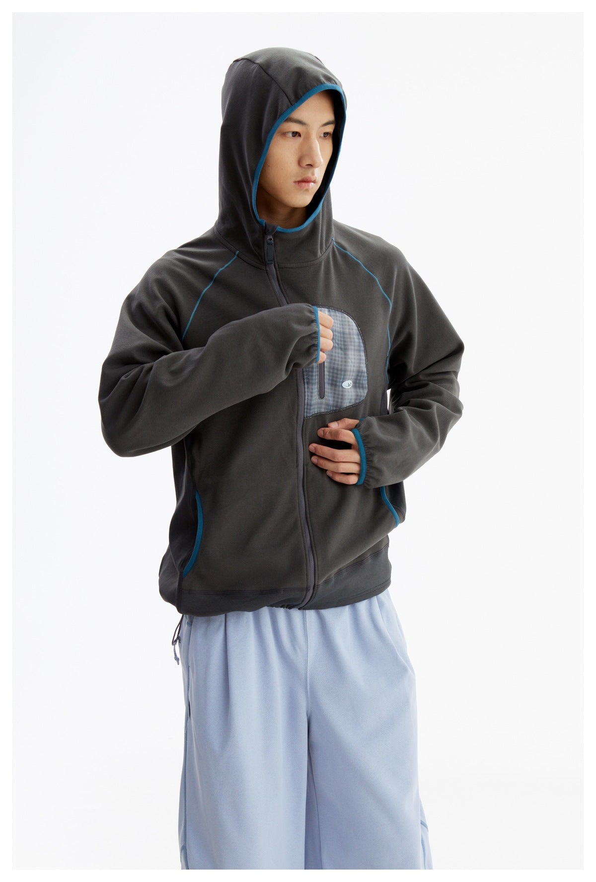 Model wearing a charcoal hooded zip-up jacket with blue trim and a plaid chest pocket, with light-blue pants.