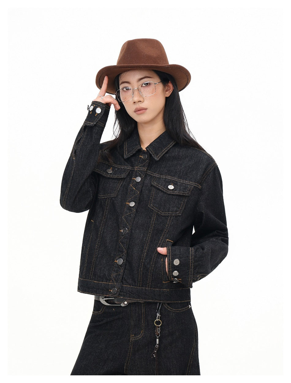 Model wearing a black denim jacket with a brown fedora and glasses, one hand in a pocket, close-up