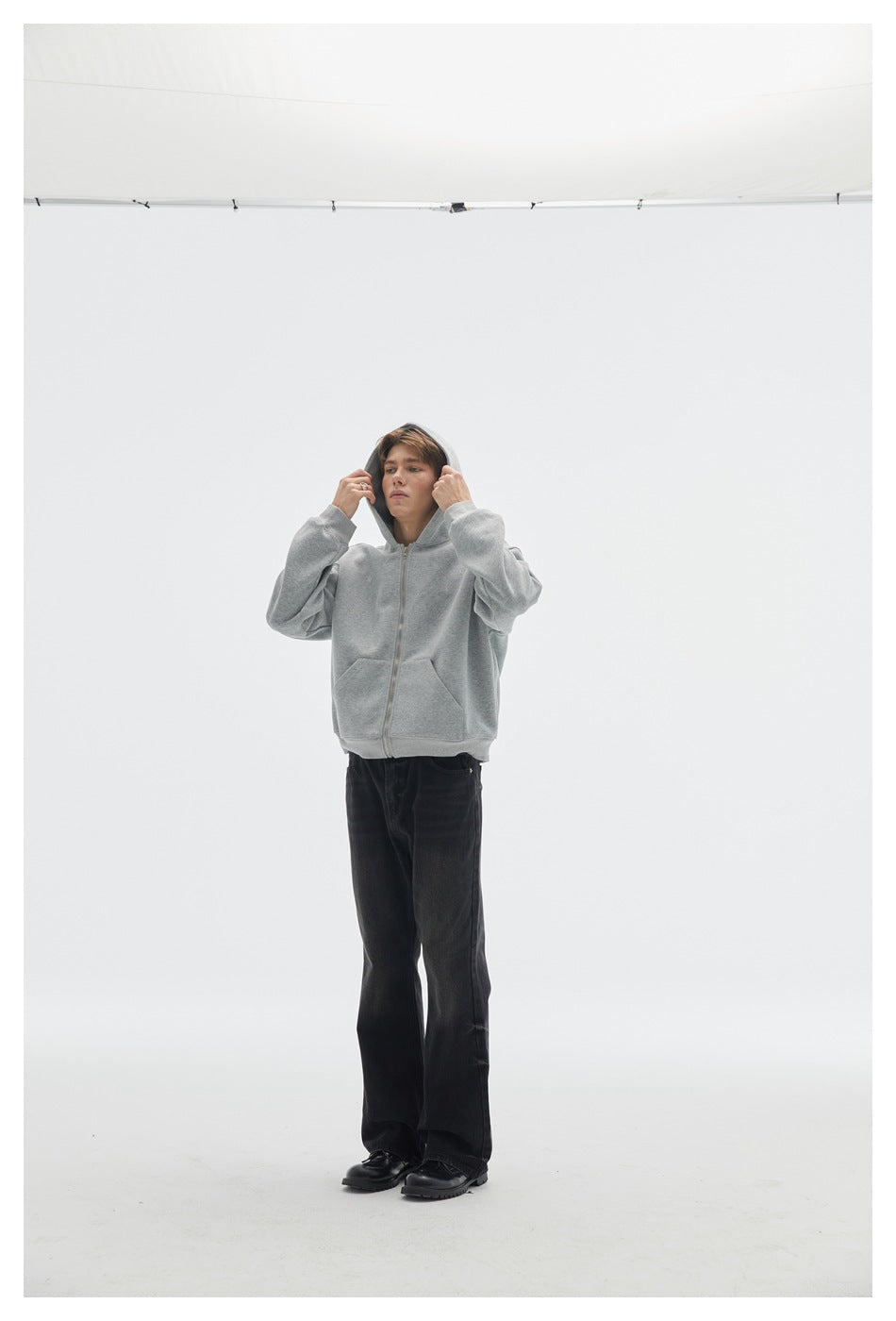 Model wearing a light gray zip hoodie with the hood raised and hands adjusting it.
