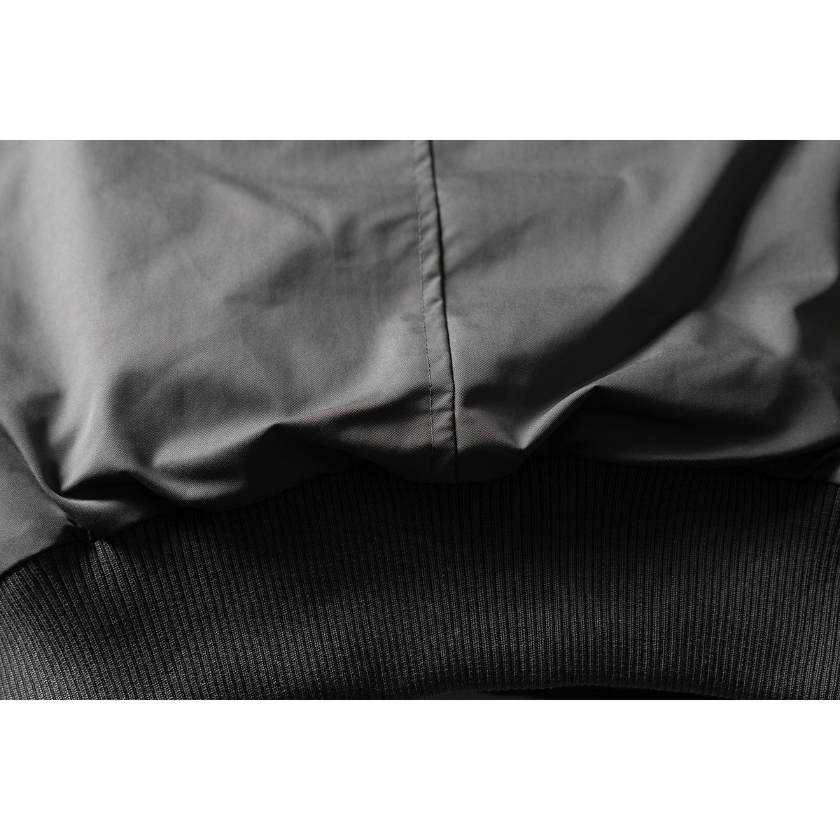 Close-up of a charcoal-black jacket’s smooth fabric, center seam, and wide black ribbed hem.