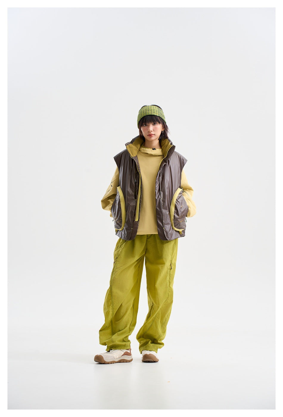 Model wearing a glossy charcoal vest with yellow-trimmed pockets, a yellow top and loose lime-green trousers.