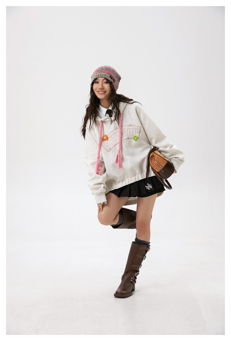 Model wearing a light gray hoodie with pink drawstrings and floral lettering, paired with a colorful knit beanie.