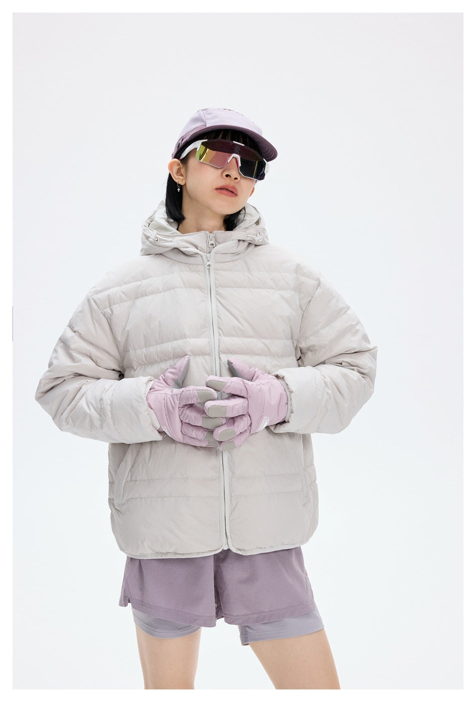 Model wearing a light gray quilted hooded jacket with a front zip, lilac gloves, shorts, cap and mirrored sunglasses.