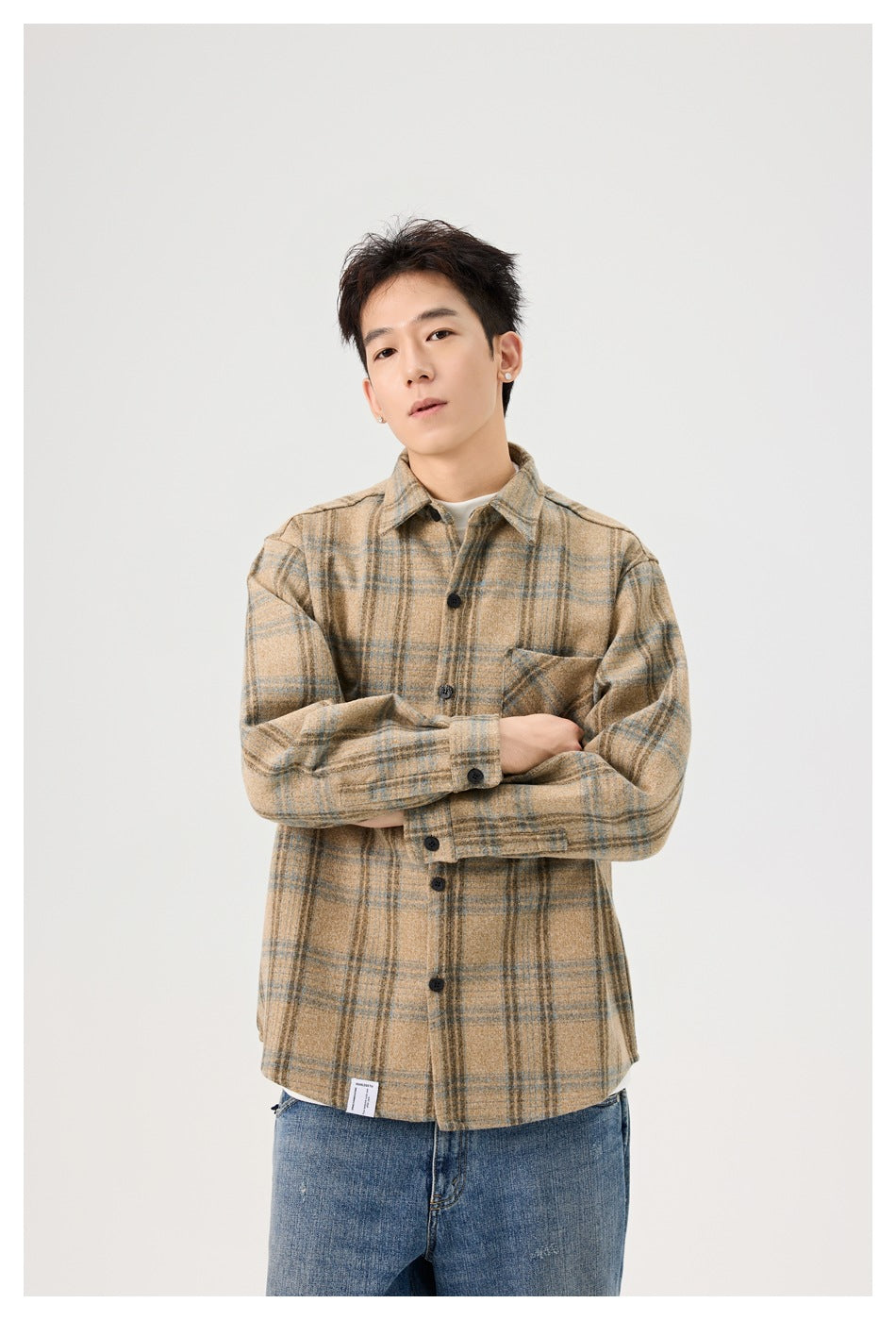 A man models a tan plaid overshirt with dark buttons and a chest pocket, arms crossed.