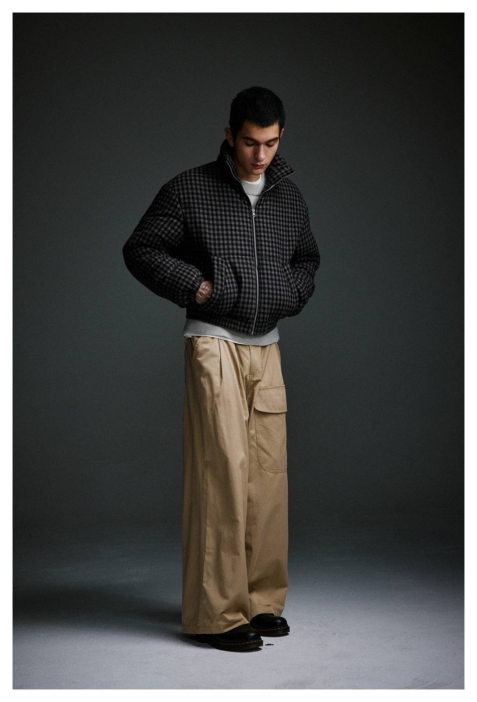 Model in a charcoal checked padded jacket and wide tan cargo trousers, looking down with hands in pockets.